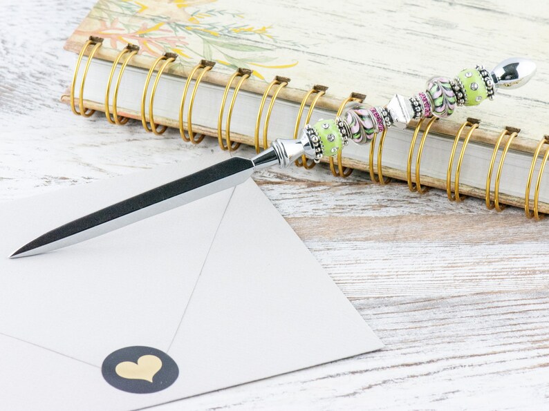 Peut inclure: Un ouvre-lettre en argent avec un manche en perles vertes et roses. Le manche est d&eacute;cor&eacute; de strass. L'ouvre-lettre repose sur un carnet ferm&eacute; avec une reliure spirale dor&eacute;e.