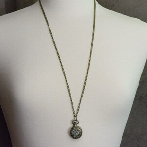 May include: A gold-toned chain necklace with a small, round pocket watch pendant. The watch face is decorated with a floral design.