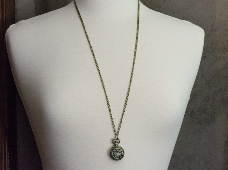 May include: A gold-toned chain necklace with a small, round pocket watch pendant. The watch face is decorated with a floral design.