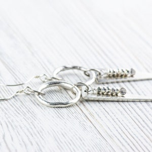 May include: A pair of silver earrings with a hammered hoop and a silver triangle pendant with a row of silver beads. The earrings are on a white wooden surface.