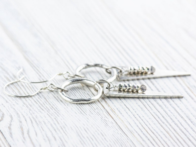 May include: A pair of silver earrings with a hammered hoop and a silver triangle pendant with a row of silver beads. The earrings are on a white wooden surface.