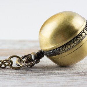 May include: A gold-toned pocket watch with a round, ornate case and a chain attached. The watch is lying on a light wood surface.
