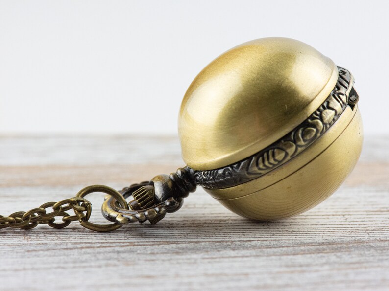 May include: A gold-toned pocket watch with a round, ornate case and a chain attached. The watch is lying on a light wood surface.