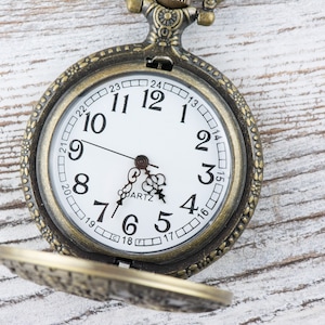 Pu&ograve; includere: Un orologio da tasca in stile vintage con quadrante bianco e indici orari romani neri. L'orologio ha una cassa in metallo dorato e un design decorativo attorno al bordo.