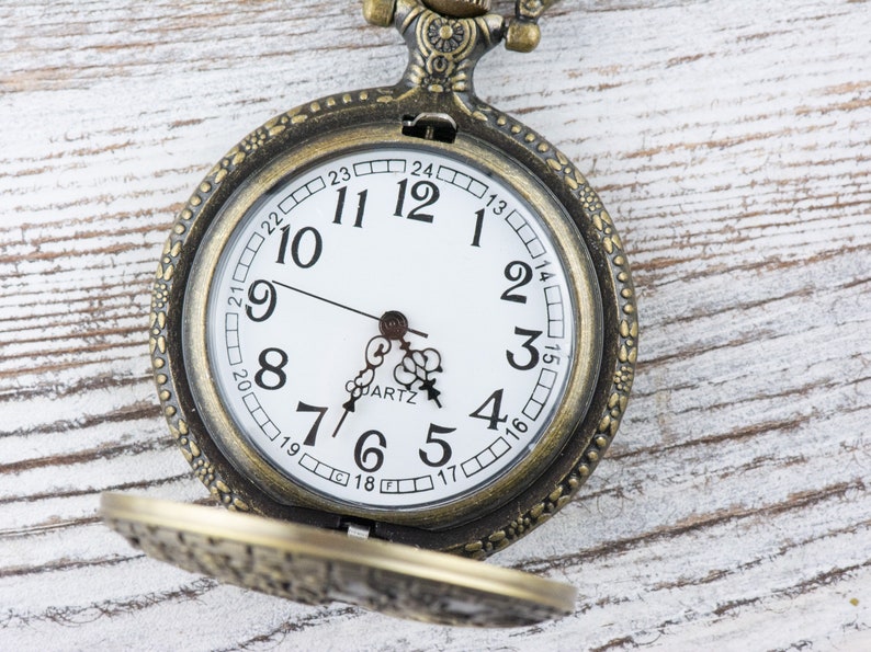 Pu&ograve; includere: Un orologio da tasca in stile vintage con quadrante bianco e indici orari romani neri. L'orologio ha una cassa in metallo dorato e un design decorativo attorno al bordo.