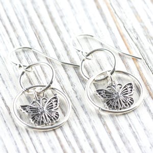 May include: A pair of silver-colored butterfly earrings. Each earring features a detailed butterfly charm suspended within a circular frame. The earrings have a hook closure and are displayed on a light-colored wooden surface.