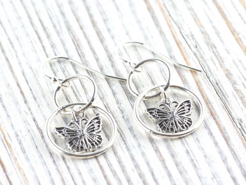 May include: A pair of silver-colored butterfly earrings. Each earring features a detailed butterfly charm suspended within a circular frame. The earrings have a hook closure and are displayed on a light-colored wooden surface.