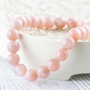 May include: A pink sunstone beaded bracelet with a smooth, polished finish. The bracelet is made of round beads and is strung on a clear elastic cord.