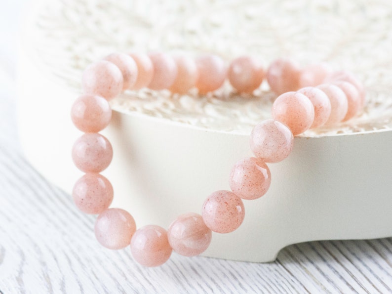 May include: A pink sunstone beaded bracelet with a smooth, polished finish. The bracelet is made of round beads and is strung on a clear elastic cord.