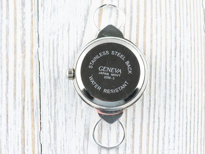Peut inclure: Le dos d'une montre argent&eacute;e avec un bo&icirc;tier rond et un fond &agrave; enclenchement. Le cadran de la montre indique "Geneva Japan Movt 2039-S" et "Stainless Steel Back Water Resistant".
