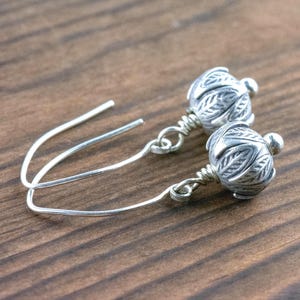 Pu&ograve; includere: Un paio di orecchini color argento con un design floreale. Gli orecchini presentano una piccola perla rotonda nella parte superiore e una sfera decorativa e strutturata nella parte inferiore. Sono esposti su una superficie di legno.