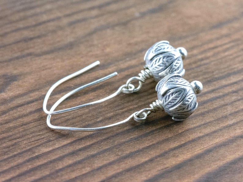 Pu&ograve; includere: Un paio di orecchini color argento con un design floreale. Gli orecchini presentano una piccola perla rotonda nella parte superiore e una sfera decorativa e strutturata nella parte inferiore. Sono esposti su una superficie di legno.