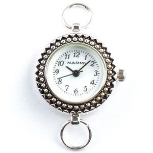 K&ouml;nnte beinhalten: Eine silberfarbene Taschenuhr mit wei&szlig;em Zifferblatt und schwarzen Zeigern. Die Uhr hat eine dekorative Perlenl&uuml;nette und einen Ring oben und unten zur Befestigung. Das Zifferblatt zeigt den Markennamen "NARMI".