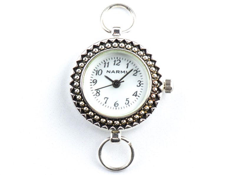 K&ouml;nnte beinhalten: Eine silberfarbene Taschenuhr mit wei&szlig;em Zifferblatt und schwarzen Zeigern. Die Uhr hat eine dekorative Perlenl&uuml;nette und einen Ring oben und unten zur Befestigung. Das Zifferblatt zeigt den Markennamen "NARMI".