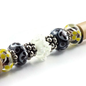 May include: A close-up of a wooden pen with black, white, and yellow glass beads. The beads have a variety of patterns, including swirls, dots, and flowers.