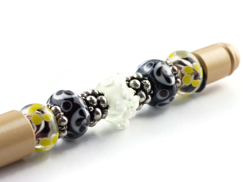 May include: A close-up of a wooden pen with black, white, and yellow glass beads. The beads have a variety of patterns, including swirls, dots, and flowers.
