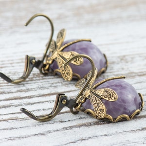 Pode incluir: Um par de brincos pendentes com uma pedra roxa e um design de lib&eacute;lula. Os brincos s&atilde;o feitos de lat&atilde;o e t&ecirc;m um fecho de gancho.