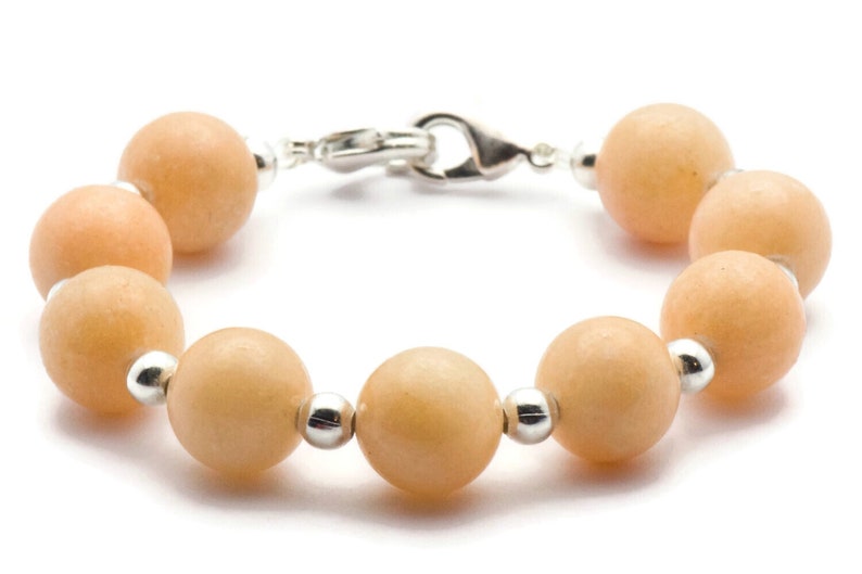 Puede incluir: Un brazalete hecho de cuentas de color naranja claro con detalles plateados. El brazalete tiene un cierre de broche.