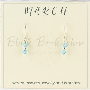 May include: A pair of sterling silver earrings with two light blue crystal beads. The earrings are displayed on a card that says "MARCH" and "Blush Brook Shop Handmade Jewelry".