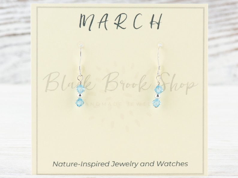 May include: A pair of sterling silver earrings with two light blue crystal beads. The earrings are displayed on a card that says "MARCH" and "Blush Brook Shop Handmade Jewelry".