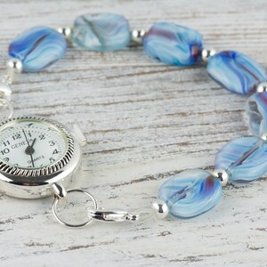 May include: A silver watch with a white face and black numbers is attached to a bracelet made of blue and white swirled glass beads.