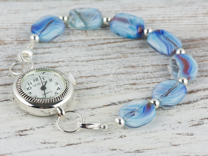 May include: A silver watch with a white face and black numbers is attached to a bracelet made of blue and white swirled glass beads.