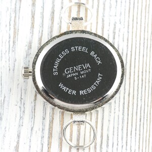 以下が含まれることがあります： 「STAINLESS STEEL BACK」、「GENEVA JAPAN MOVT S-147」、および「WATER RESISTANT」の文字が入った、銀色の時計の裏側のクローズアップ。 時計は丸い文字盤と小さなリューズが付いています。 背景は白い木製の表面です。