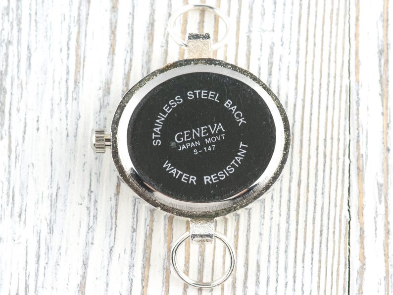 以下が含まれることがあります： 「STAINLESS STEEL BACK」、「GENEVA JAPAN MOVT S-147」、および「WATER RESISTANT」の文字が入った、銀色の時計の裏側のクローズアップ。 時計は丸い文字盤と小さなリューズが付いています。 背景は白い木製の表面です。