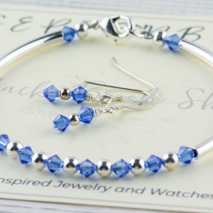 May include: A silver bangle bracelet with a row of blue crystal beads and matching blue crystal dangle earrings. The bracelet and earrings are on a white background with the text "Black Bead Shop Handmade Jewellery".