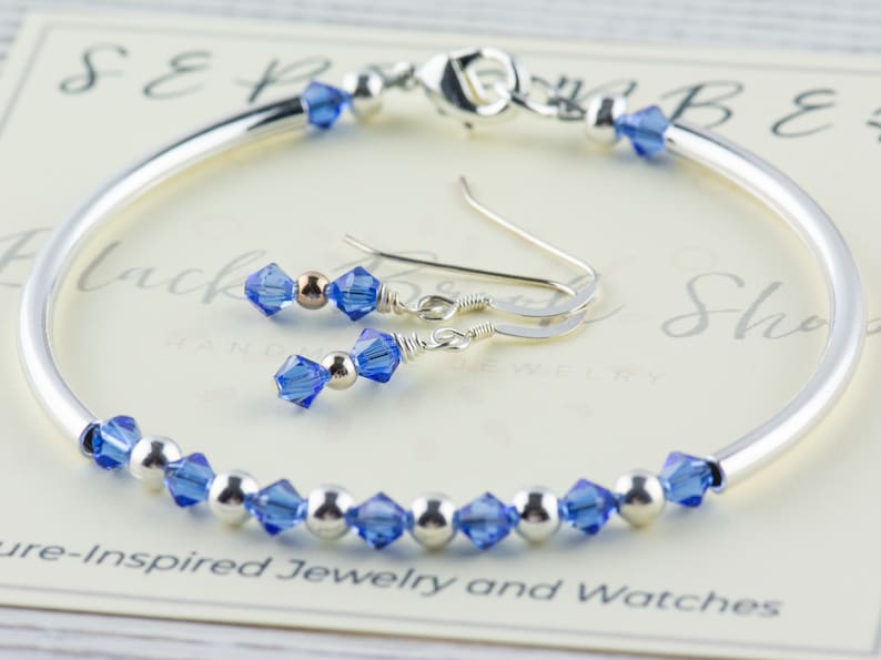 May include: A silver bangle bracelet with a row of blue crystal beads and matching blue crystal dangle earrings. The bracelet and earrings are on a white background with the text "Black Bead Shop Handmade Jewellery".