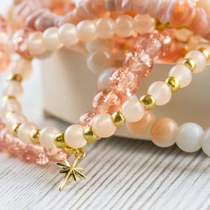 May include: A set of layered bracelets with pink and white beads and gold accents. The bracelets have a star charm attached.