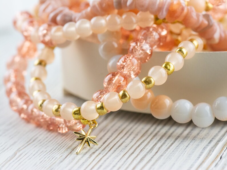 May include: A set of layered bracelets with pink and white beads and gold accents. The bracelets have a star charm attached.