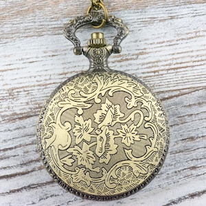 Pu&ograve; includere: Un orologio da tasca dorato con un design floreale sul davanti. L'orologio ha una catena attaccata alla parte superiore.