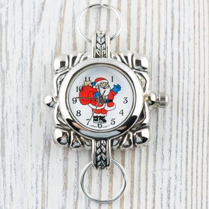 May include: Silver-toned pocket watch with a white face featuring a cartoon Santa Claus holding a red bag. The watch has a square face with a decorative border and a key ring attached.