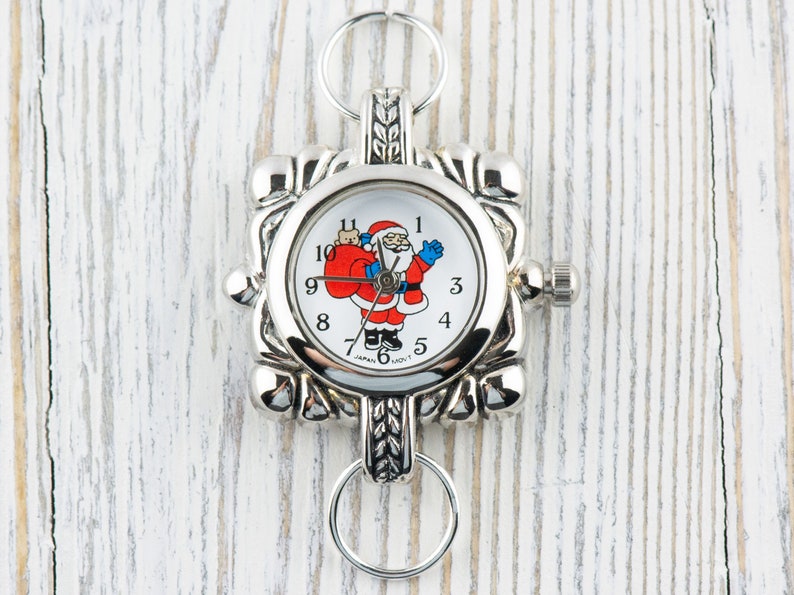 May include: Silver-toned pocket watch with a white face featuring a cartoon Santa Claus holding a red bag. The watch has a square face with a decorative border and a key ring attached.