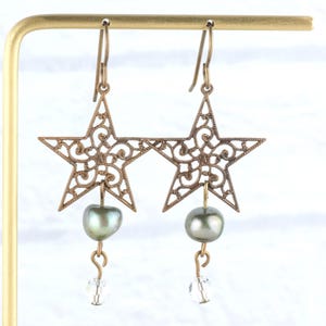 May include: A pair of bronze-colored star-shaped earrings with intricate filigree detailing. Each earring features a dark green pearl and a clear crystal bead. The earrings are displayed on a gold-colored stand.
