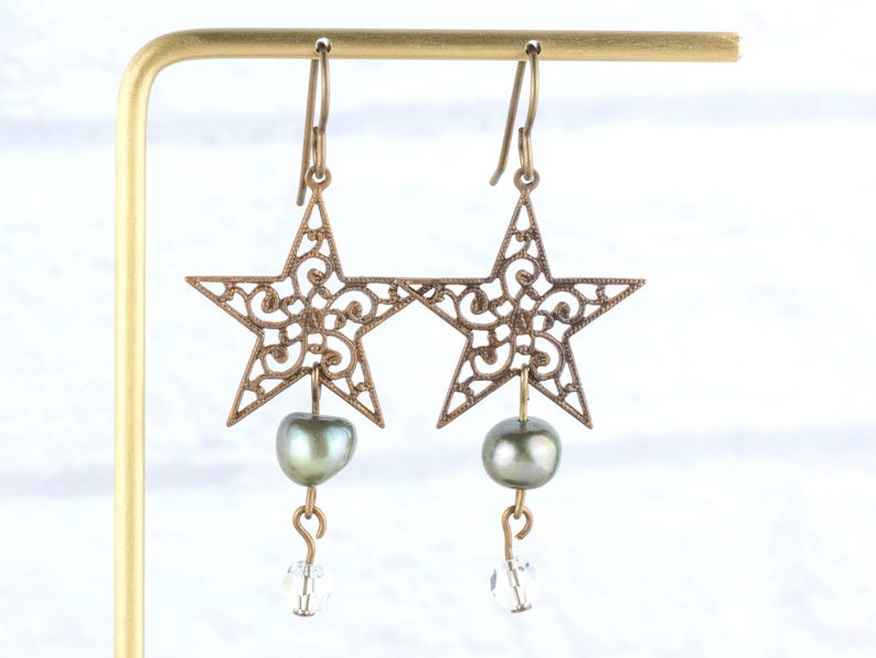 May include: A pair of bronze-colored star-shaped earrings with intricate filigree detailing. Each earring features a dark green pearl and a clear crystal bead. The earrings are displayed on a gold-colored stand.