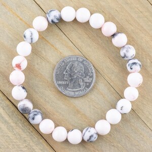May include: A pink and white gemstone bracelet with round beads. The bracelet is laid out on a wooden surface next to a US quarter for size reference.