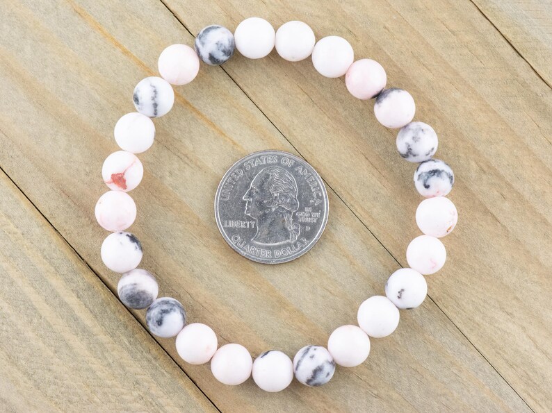 May include: A pink and white gemstone bracelet with round beads. The bracelet is laid out on a wooden surface next to a US quarter for size reference.