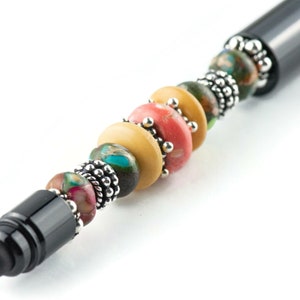 K&ouml;nnte beinhalten: Ein schwarzer Stift mit einem dekorativen Perlenkopf. Die Perlen sind mehrfarbig und umfassen Farbt&ouml;ne von Gr&uuml;n, Gelb, Rosa und Braun. Die Perlen sind durch silberne Metallabstandshalter getrennt.