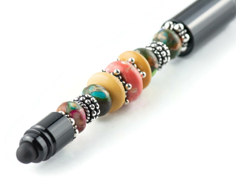 K&ouml;nnte beinhalten: Ein schwarzer Stift mit einem dekorativen Perlenkopf. Die Perlen sind mehrfarbig und umfassen Farbt&ouml;ne von Gr&uuml;n, Gelb, Rosa und Braun. Die Perlen sind durch silberne Metallabstandshalter getrennt.