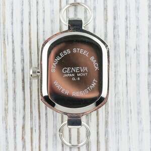 May include: A close-up of the back of a Geneva watch, featuring a stainless steel back and water-resistant design. The watch has a rectangular shape with a silver-tone metal frame and two loops. The text "Geneva Japan Movt CL-8" is visible.