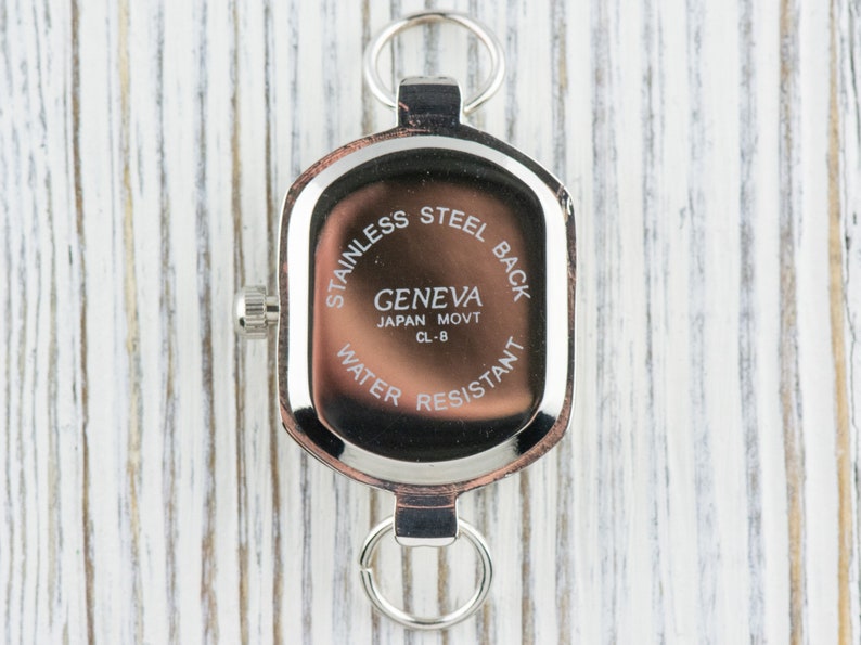 May include: A close-up of the back of a Geneva watch, featuring a stainless steel back and water-resistant design. The watch has a rectangular shape with a silver-tone metal frame and two loops. The text "Geneva Japan Movt CL-8" is visible.