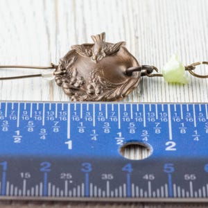 May include: An antique bronze earring with a decorative circular pendant featuring a bird and floral design. The earring includes a clear crystal bead and a light green bead. The earring measures approximately 2 inches in length.