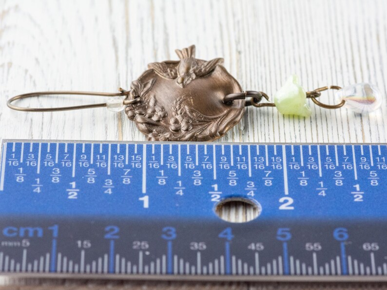 May include: An antique bronze earring with a decorative circular pendant featuring a bird and floral design. The earring includes a clear crystal bead and a light green bead. The earring measures approximately 2 inches in length.