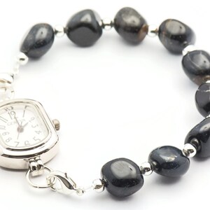 K&ouml;nnte beinhalten: Eine silberne Uhr mit quadratischem Zifferblatt und schwarzen Zahlen ist an einem Armband aus schwarzen Edelsteinperlen befestigt.