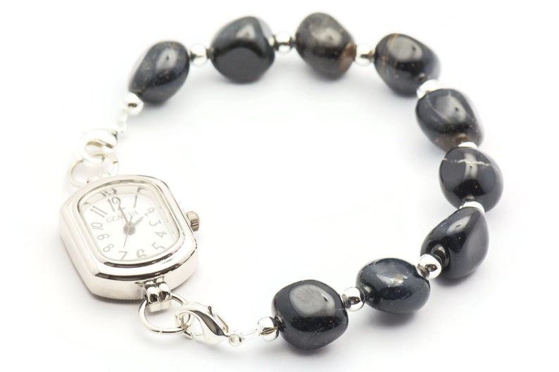 K&ouml;nnte beinhalten: Eine silberne Uhr mit quadratischem Zifferblatt und schwarzen Zahlen ist an einem Armband aus schwarzen Edelsteinperlen befestigt.