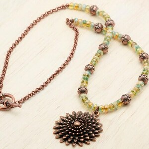 Pu&ograve; includere: Una collana artigianale con una catena color rame e un ciondolo a forma di fiore. La collana ha una chiusura a toggle ed &egrave; adornata con perline verdi e gialle e perline decorative in rame.