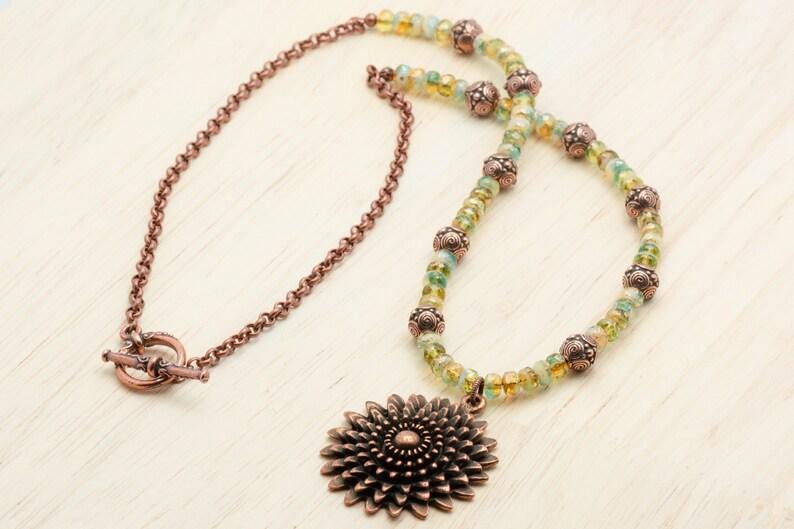 Pu&ograve; includere: Una collana artigianale con una catena color rame e un ciondolo a forma di fiore. La collana ha una chiusura a toggle ed &egrave; adornata con perline verdi e gialle e perline decorative in rame.
