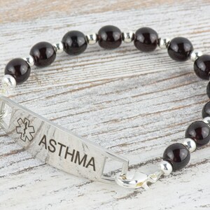 K&ouml;nnte beinhalten: Ein silbernes medizinisches Armband mit schwarzen Perlen und dem Wort "ASTHMA" auf dem Anh&auml;nger. Der Anh&auml;nger zeigt auch ein medizinisches Symbol.